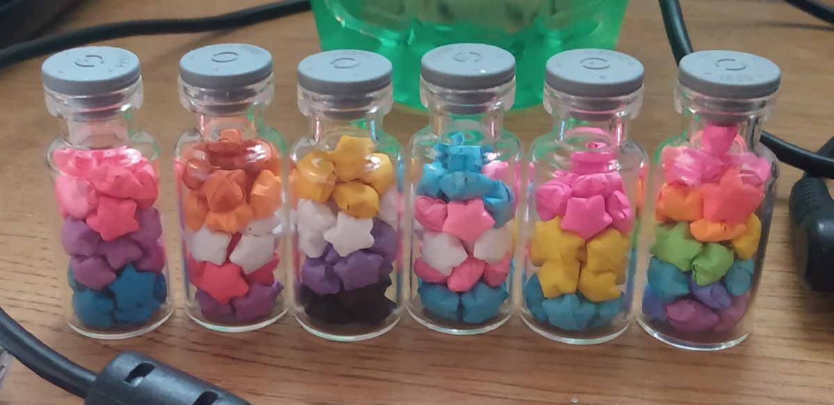 multiple vials filled with colored stars. the colors make pride flags in the following order: bi, lesbian, nonbinary, trans, pan, and rainbow