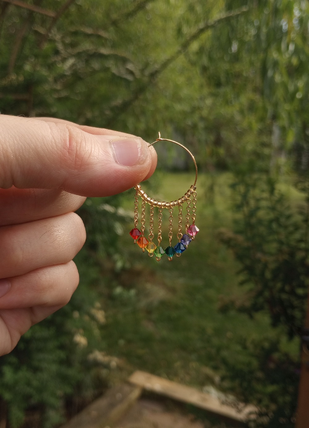 close up of a hoop earing worn by me. eight beads dangle from the hoop by thin gold chains, each one a color of the rainbow.