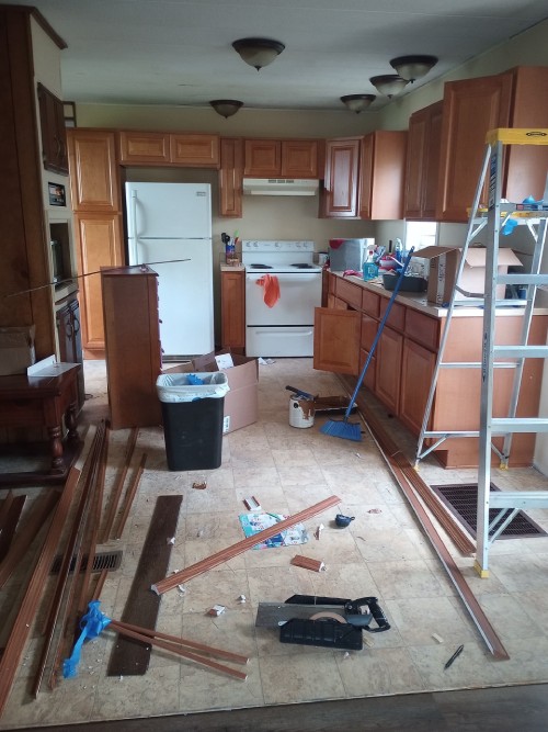 A small messy kitchen with trim and tools all over the floor and counters. A ladder stands to the side.