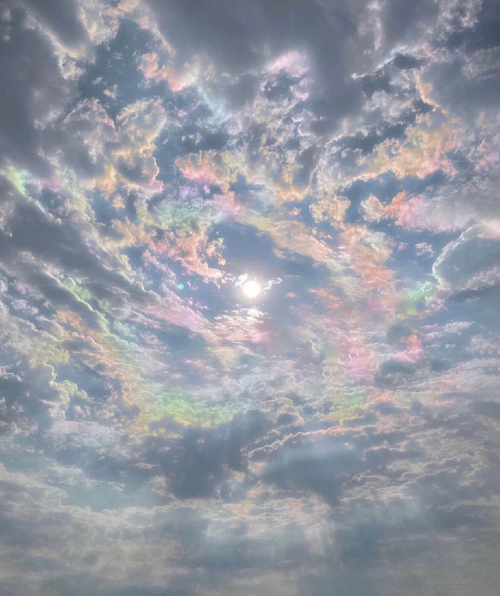 sky full of patchy thin clouds surrounding the sun. Patches of pastel rainbow colors glint off of the clouds.