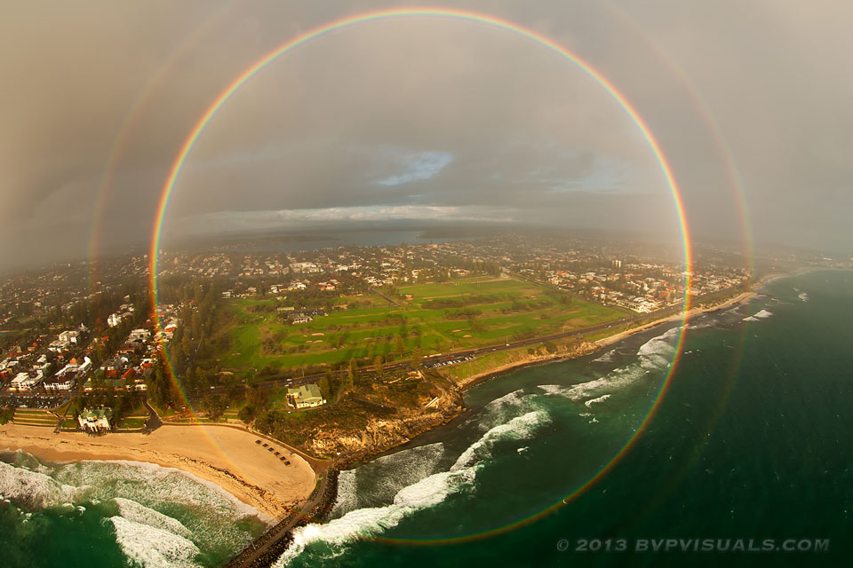 A full rainbow and faint secondary rainbow, viewed against a shoreline populated by buildings.
