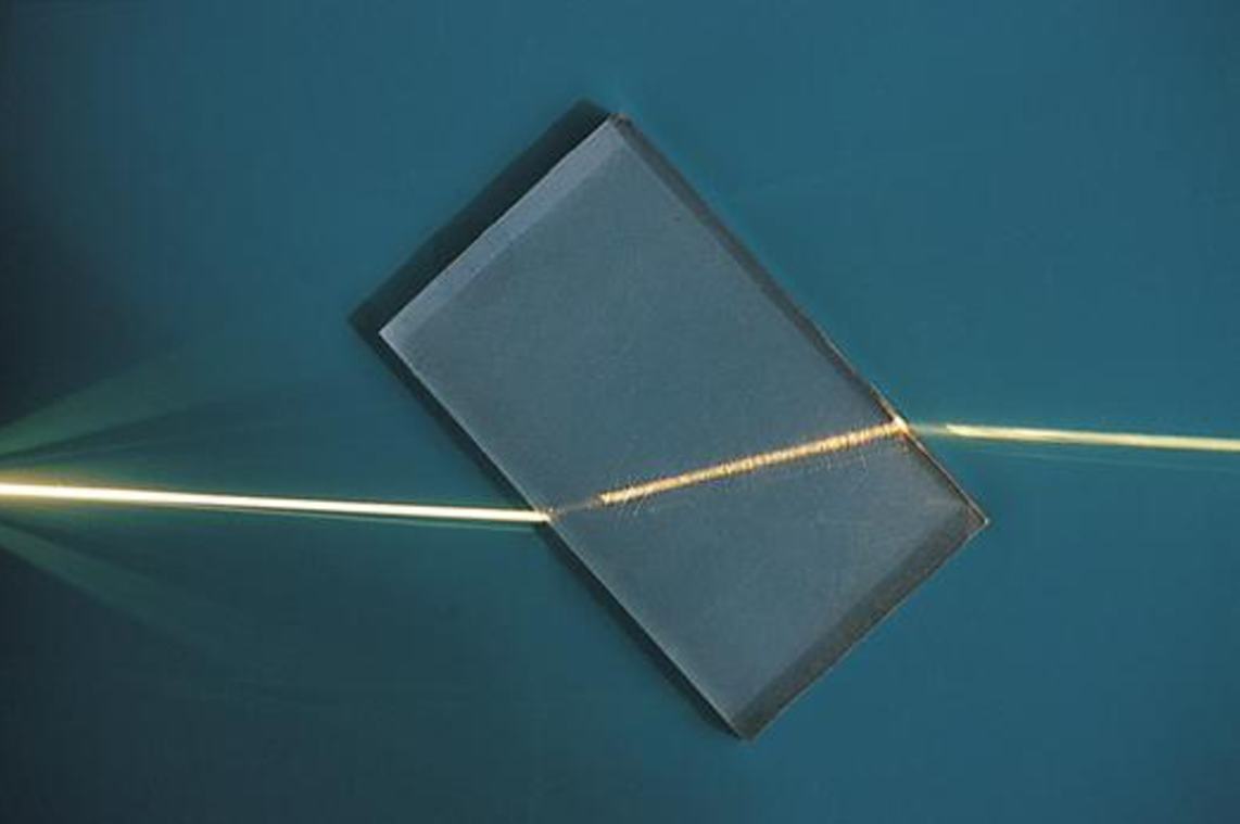photo of yellow lasar light passing through a block of glass at an angle. The light tilts at a different angle inside the glass.