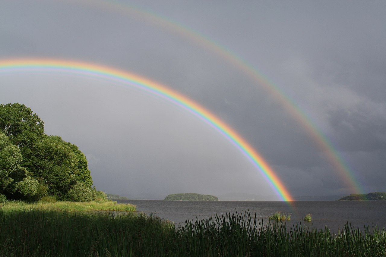a bright primary and faint secondary rainbow over a body of water and trees